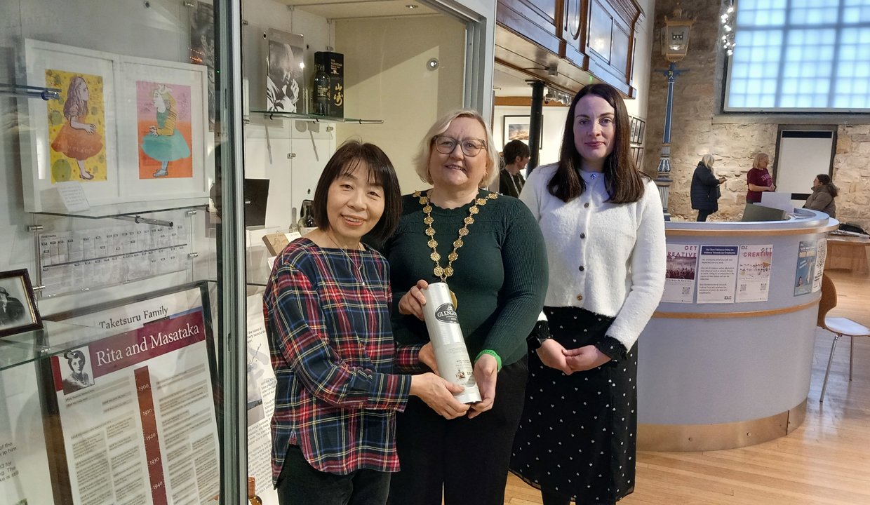 Standing next to the Rita and Masataka display in the Auld Kirk Museum: Naoko Nakamato and Provost Gillian Renwick hold a commemorative bottle of whisky presented to Naoko, with Depute Provost Colette McDiarmid standing nearby